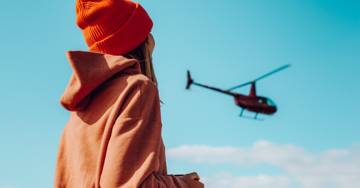 <img src="Moda-bagażowa"alt="dziewczyna w w pomarańczowej bluzie i czapce patrząca na odlatujący helikopter">