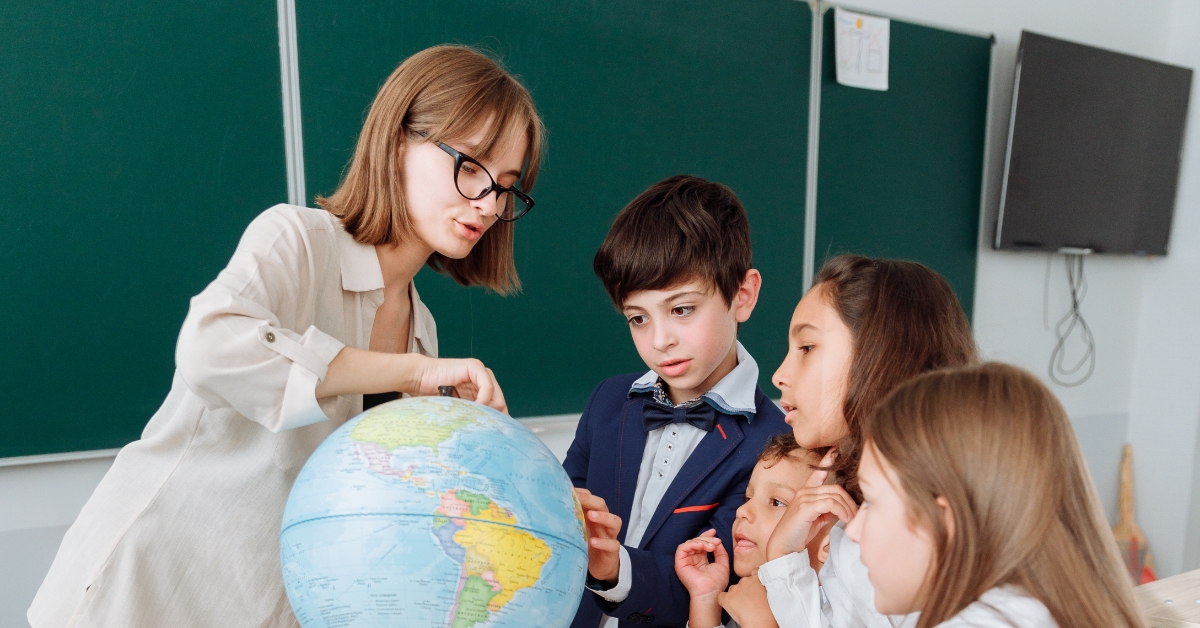 <img src=”wyprawka-szkolna"alt="Nauczycielka pokazująca globus uczniom w klasie">